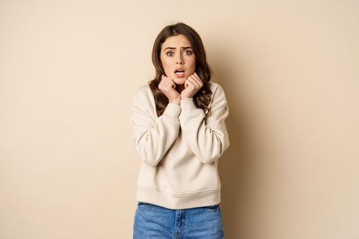 Scared insecure woman trembling from fear, looking at camera shocked and frightened, standing over beige background.