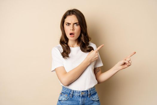 Frustrated woman looking insulted, pointing fingers right and gasping shocked, standing in t-shirt over beige background.
