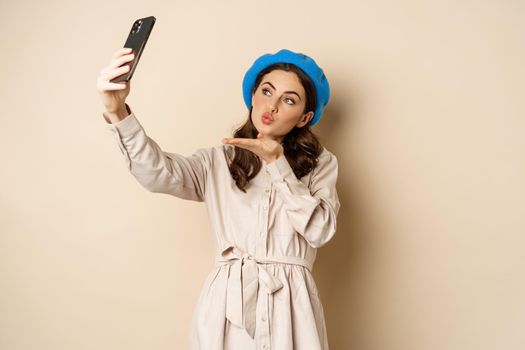 Beautiful modern woman posing for selfie, holding smartphone, taking picture photo on mobile phone, standing happy against beige background.