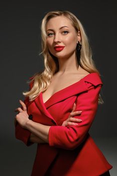 Beautiful blonde girl with bright make-up and deep neckline, hands folded, in red dress and black earrings. She smiling, posing sideways on gray background. Fashion and beauty. Copy space, close up