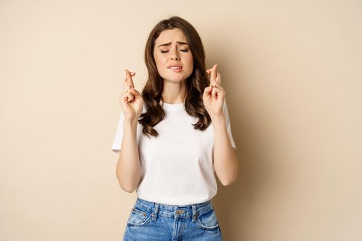 Hopeful woman praying, cross fingers for good luck, making wish, standing over beige background.