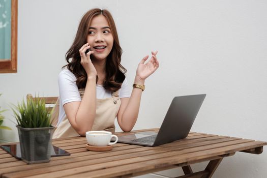 Smiling young woman talking on the phone while using laptop at cafe shop, happy young girl holds cellphone making answering call, attractive teenager having pleasant conversation chatting by mobile with friend.