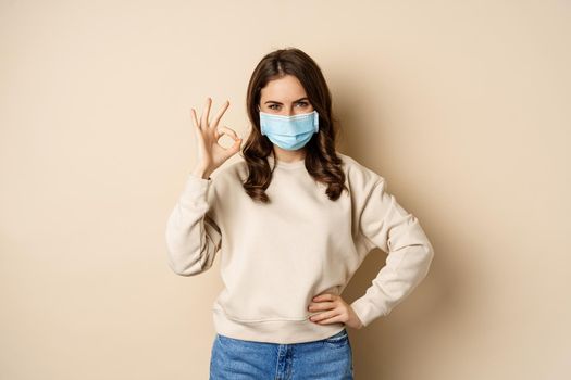 Covid-19, pandemic and quarantine concept. Young woman wears medical face mask during coronavirus omicron outbreak, showing okay sign, standing over beige background.