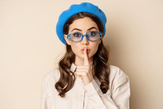 Secret. Young cute woman in sunglasses, shushing, showing taboo, hush sign, press finger to lips, standing over beige background.