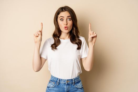 Excited girl looking interested, pointing fingers up, showing advertisement, standing in tshirt over beige background.