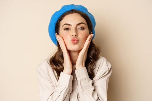 Image of stylish modern girl with makeup, clear glowing skin, pucker lips kissing face, standing over beige background coquettish.