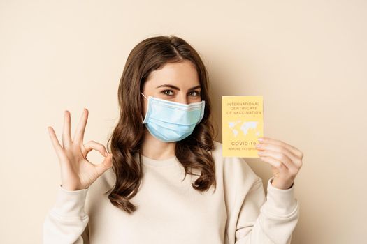 Covid and people concept. Vaccinated woman in face mask showing covid international vaccination certificate, recommend get vaccine, standing over beige background.