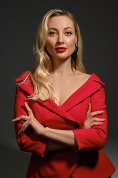 Gorgeous blonde female with bright make-up and deep neckline, hands folded, in red dress and black earrings. She is smiling, posing against gray background. Fashion and beauty. Copy space, close up