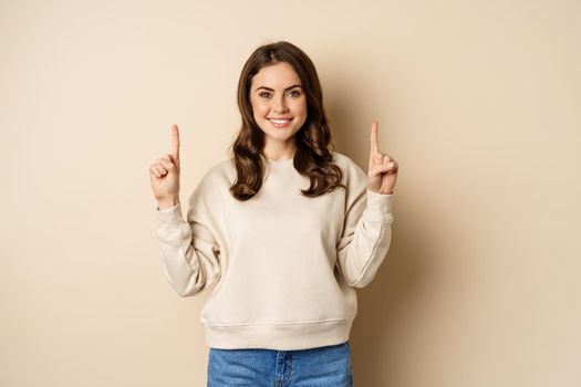 Enthusiastic girl pointing fingers up, showing announcement, promo text or logo upwards, standing over beige background.