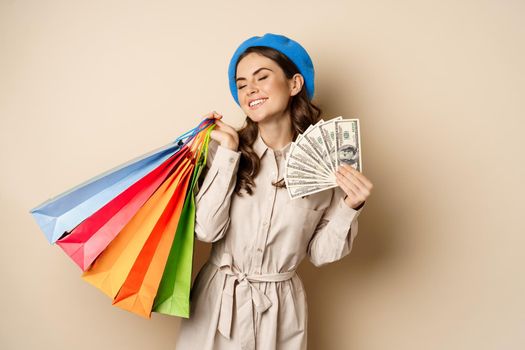 Stylish modern girl feeling satisfaction while shopping, posing with money dollars and shop bags, beige studio background.