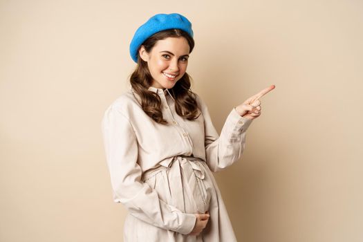 Young future mother, pregnant woman smiling, posing with big belly third trimester of pregnancy, looking happy, expecting baby, pointing right at logo, banner or advertisement.