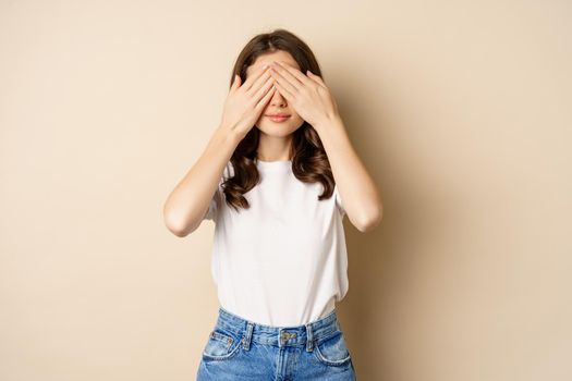 Surprise. Happy young woman shut eyes with hands and waiting for gift, standing over beige background.