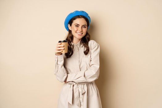 Beautiful modern girl posing with takeout cup of coffee, drinking from reusable cup from cafe and posing happy, beige studio background.