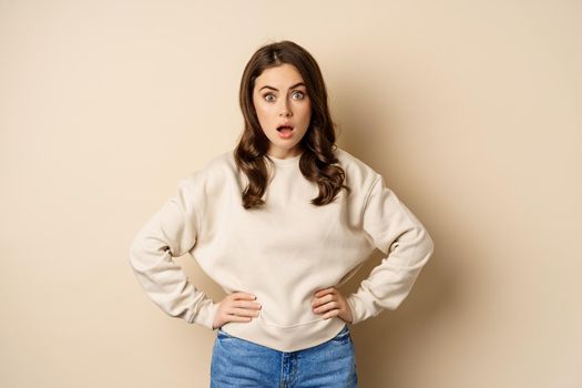 Portrait of shocked brunette woman drop jaw, gasping and staring speechless at camera, beige background. Copy space