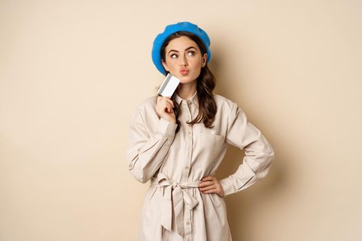 Shopping and discounts concept. Young stylish modern girl, posing with credit card, looking happy and smiling, standing over beige background.