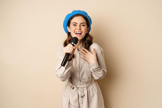 Portrait of happy stylish woman performing, singing with microphone, posing against beige background.