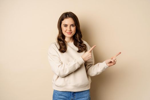 Smiling gorgeous woman pointing fingers right, inviting people, showing logo or banner, standing over beige background.