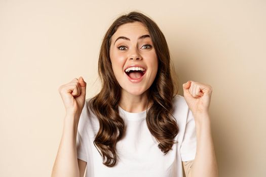 Close up of cheerful, happy beautiful woman winning, chanting and rejoicing, shouting supportive and looking hopeful at camera, yearning to win, beige background.