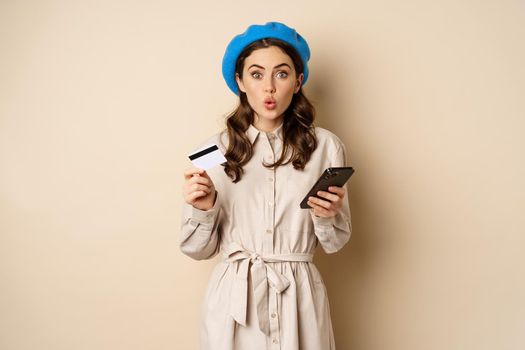 Portrait of girl looking excited, say wow, holding credit card and mobile phone, standing over beige background. Copy space