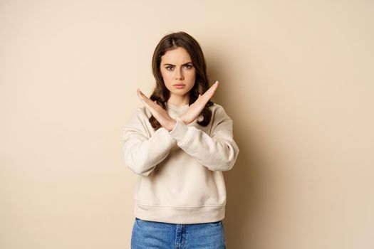 Woman showing stop, cross sign gesture and frowning angry, dislike and disapprove, saying no, rejecting and prohibiting, standing over beige background.