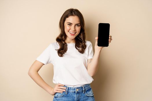 Online shopping and app concept. Young woman smiling and showing mobile phone screen, application interface, standing over beige background.