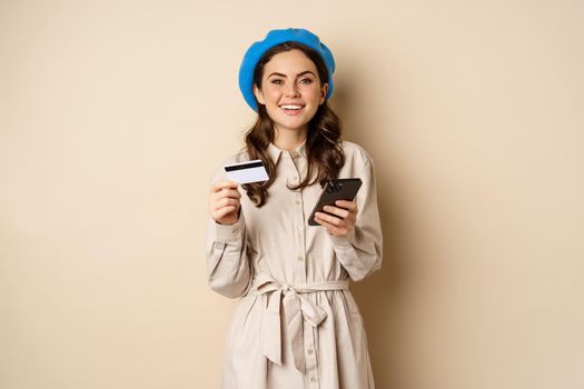 Portrait of beautiful young woman, 25 years old, using mobile phone shopping app, credit card, order delivery, purchase, standing over beige background.