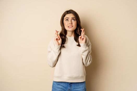 Hopeful worried girl cross fingers, making wish, standing over beige background.