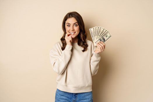 Excited brunette woman holding money and biting finger with yearning face, watns to buy smth, shopoholic with cash, standing over beige background.