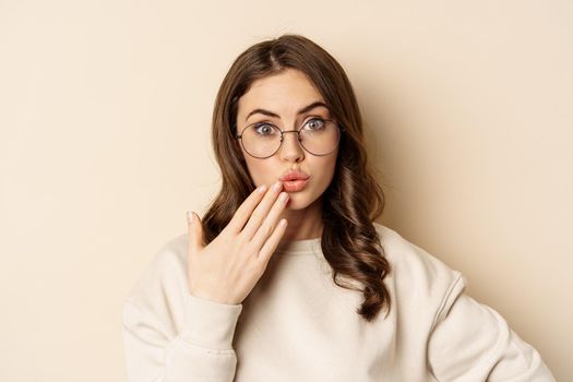 Close up portrait of girl in glasses looking surprised, say oops, standing coquettish over beige background. Copy space