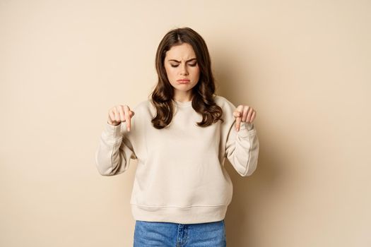 Sad and disappointed girl pointing, looking down with upset grimace moody face, standing over beige background.