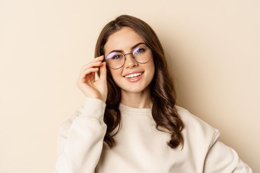 Close up portrait of beautiful modern woman in glasses, smiling and looking happy, posing in eyewear against beige background.