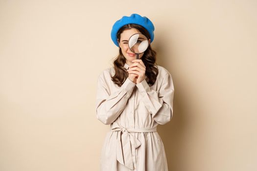 Concept of search. Young stylish girl in detective clothing, looking through magnifying glass, big eye, searching, looking for something, standing over beige background.