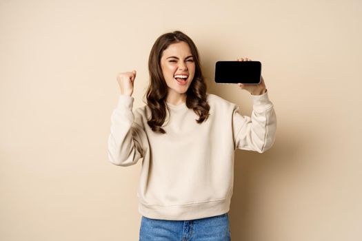 Happy girl showing mobile phone horizontal screen and celebrating, chanting with joyful face, standing over beige background.