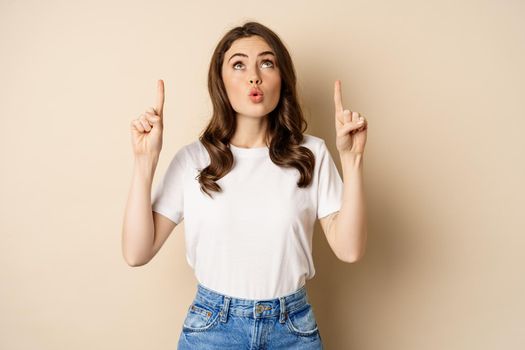 Surprised brunette girl showing promo, pointing and looking up with amazed face expression, standing over beige background.