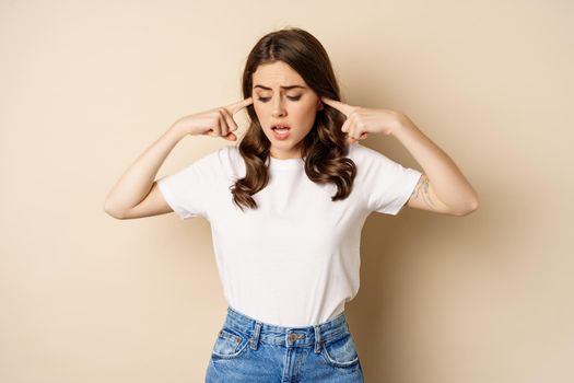 Annoyed young woman shut ears from loud noise, feeling discomfort and complaining, standing against beige background.