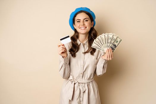 Microcredit and money concept. Young stylish woman showing credit card and dollars cash, smiling happy and satisfied, standing over beige background.