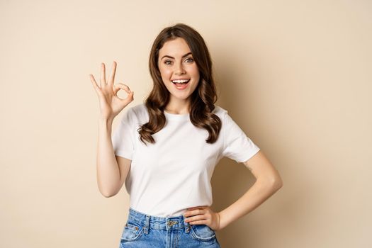 Beautiful female model smiling pleased, showing okay, zero sign, complimenting, praising you, standing over beige background.