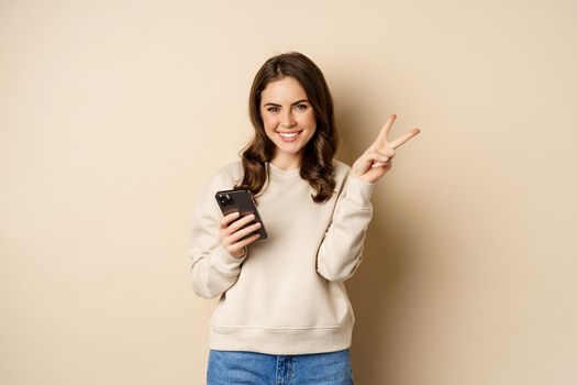 Beautiful smiling woman showing peace sign, holding mobile phone, standing over beige background. Copy space