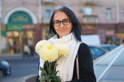 A beautiful middle-aged woman getting grey-haired in a dark coat in a spring town with a bouquet of flowers