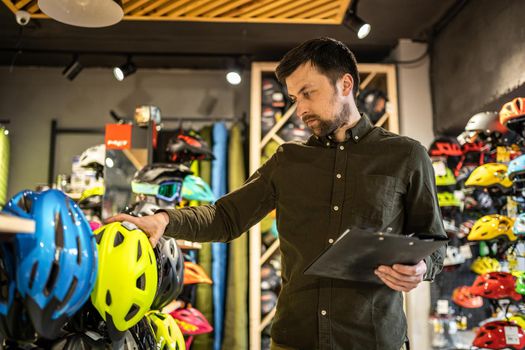 A male bike shop manager makes an inventory of sports helmets in a bike shop. The owner of a sports store with a clipboard in his hands checks the prices of bicycle helmets in the showcase.