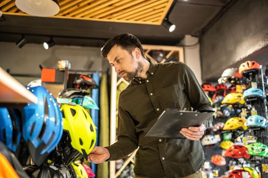 Bike shop manager checks helmet price information on tablet, seller makes an inventory in sports shop. Theme of small business selling bicycles. Seller in hands document checklist in bicycle store.