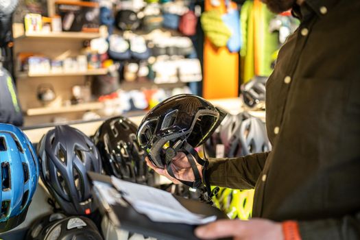 A male bike shop manager makes an inventory of sports helmets in a bike shop. The owner of a sports store with a clipboard in his hands checks the prices of bicycle helmets in the showcase.