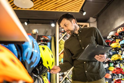 Bike shop manager checks helmet price information on tablet, seller makes an inventory in sports shop. Theme of small business selling bicycles. Seller in hands document checklist in bicycle store.