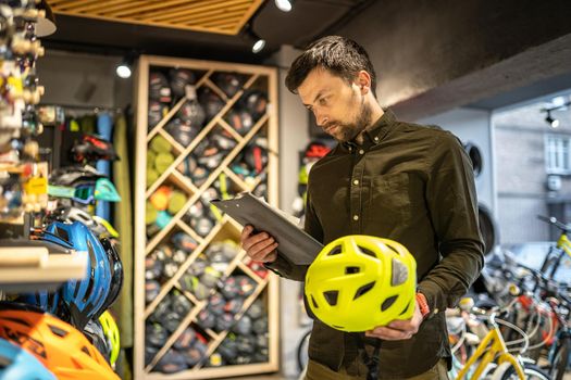 A male bike shop manager makes an inventory of sports helmets in a bike shop. The owner of a sports store with a clipboard in his hands checks the prices of bicycle helmets in the showcase.