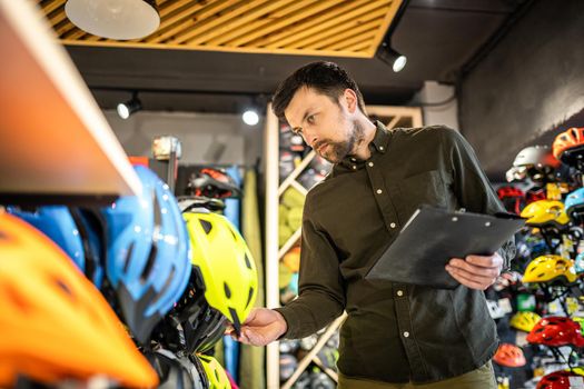 A male bike shop manager makes an inventory of sports helmets in a bike shop. The owner of a sports store with a clipboard in his hands checks the prices of bicycle helmets in the showcase.