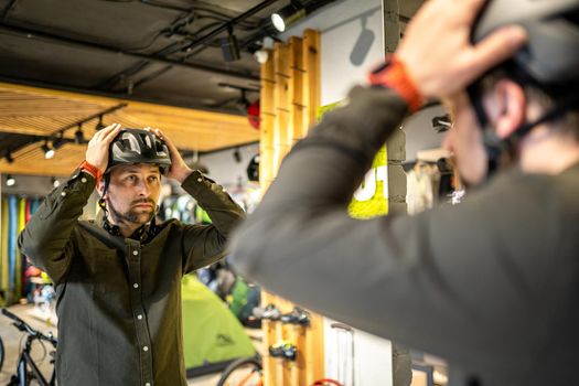 Caucasian man trying on black bicycle helmet near mirror in sporting goods store. Male buyer chooses safety helmet for cycling. Shopping in bicycle store. Person adjusts his sports helmet strap.