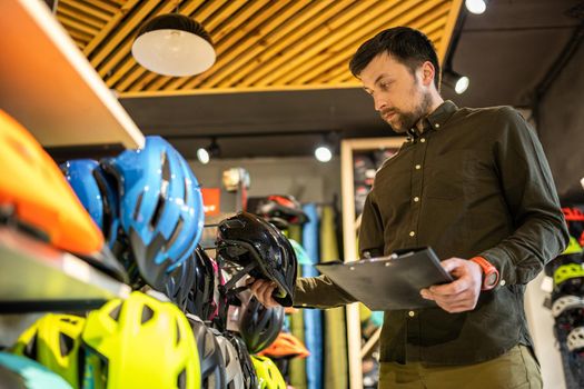 A male bike shop manager makes an inventory of sports helmets in a bike shop. The owner of a sports store with a clipboard in his hands checks the prices of bicycle helmets in the showcase.