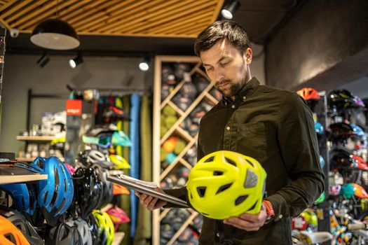 Bike shop manager checks helmet price information on tablet, seller makes an inventory in sports shop. Theme of small business selling bicycles. Seller in hands document checklist in bicycle store.