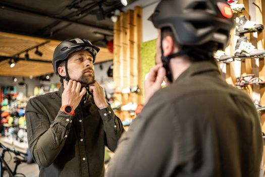 Young man came to the bicycle store. He is measuring the helmet. Male chooses helmet in sports equipment store. Purchase of new sports helmet. Customer with bicycle helmet trying on near the mirror.