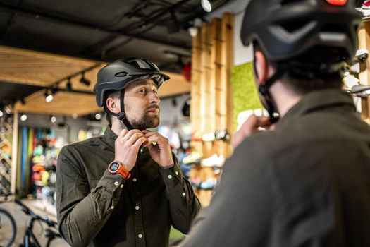 Young man came to the bicycle store. He is measuring the helmet. Male chooses helmet in sports equipment store. Purchase of new sports helmet. Customer with bicycle helmet trying on near the mirror.